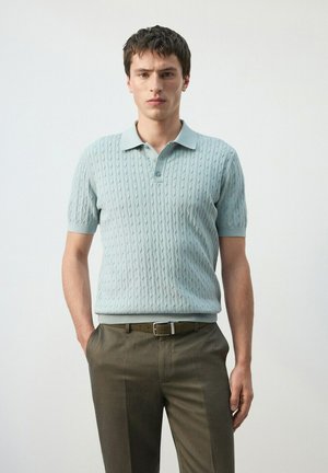 Giovane uomo in piedi con le mani in tasca, indossa una polo a maniche corte azzurro chiaro lavorata a trecce e pantaloni verde oliva con cintura.