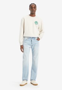 Light gray sweatshirt with a round neckline and green logo on the chest, paired with light blue straight-leg jeans and white sneakers.