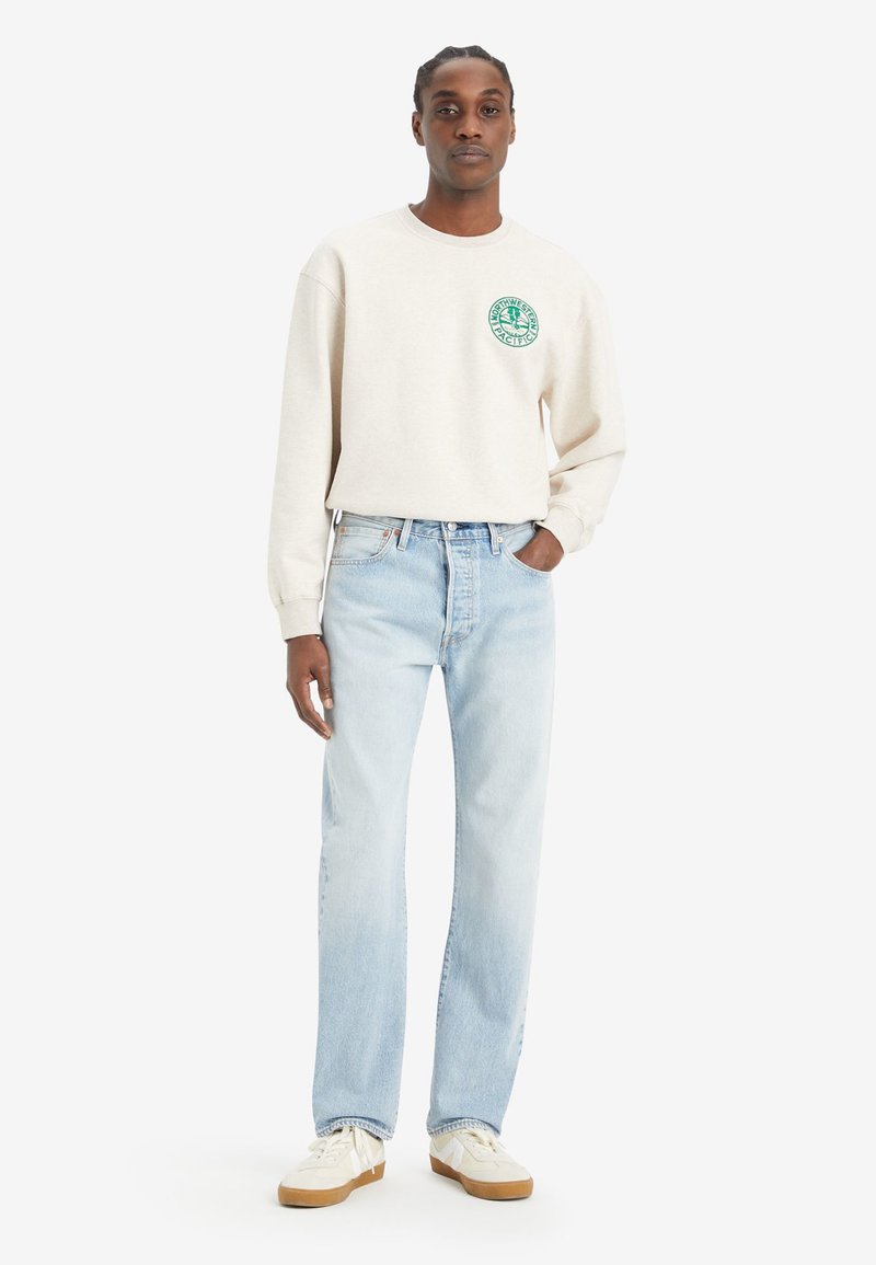 Light gray sweatshirt with a round neckline and green logo on the chest, paired with light blue straight-leg jeans and white sneakers.