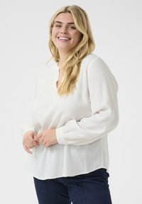 Bílá blouse s dlouhým rukávem a texturovaným vzorem, kulatým výstřihem a elastickými manžetami, v kombinaci s tmavými džínami.