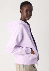 Šviesiai violetinė zip-up pūkinė striukė su erdvia kapišonu, priekiniame kišenėse ir gumbuotais rankogaliais. Minkšta tekstūra ir kasdieniška dizainas, vilkima ant džinsų.