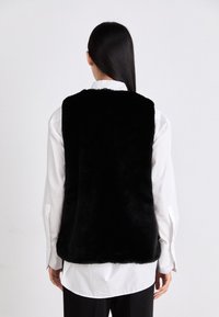 Schwarzige Kunstfellweste mit weicher, plüschiger Textur. Sie hat ein ärmelloses Design und einen abgerundeten Saum und wird über einem weißen Langarmshirt getragen.