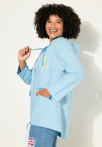 Chaqueta con capucha de color azul claro y cremallera frontal, con un gráfico de carita sonriente en amarillo, puños elásticos y dobladillo ajustable. Acompañada de jeans desgastados.