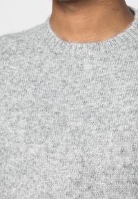 Primo piano di una persona che indossa un maglione lavorato a maglia grigio chiaro con un colletto a girocollo a coste.