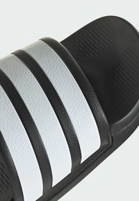 Sandale noire à glisser avec trois rayures blanches texturées sur le dessus. Le lit de pied a une finition lisse avec une semelle extérieure à motif pour l'adhérence.