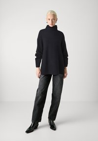 Temno pletena turtleneck pulover z dolgimi rokavi, v kombinaciji z ohlapnimi črnimi kavbojkami iz denima in črnimi čevlji s peto. 