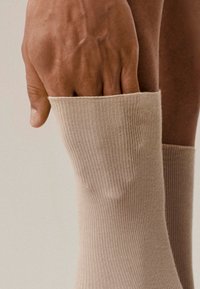 Beżowe skarpety prążkowane o miękkiej fakturze, z wywiniętym mankietem i gładkim wykończeniem, trzymane w dłoni ukazującej subtelne detale skóry.