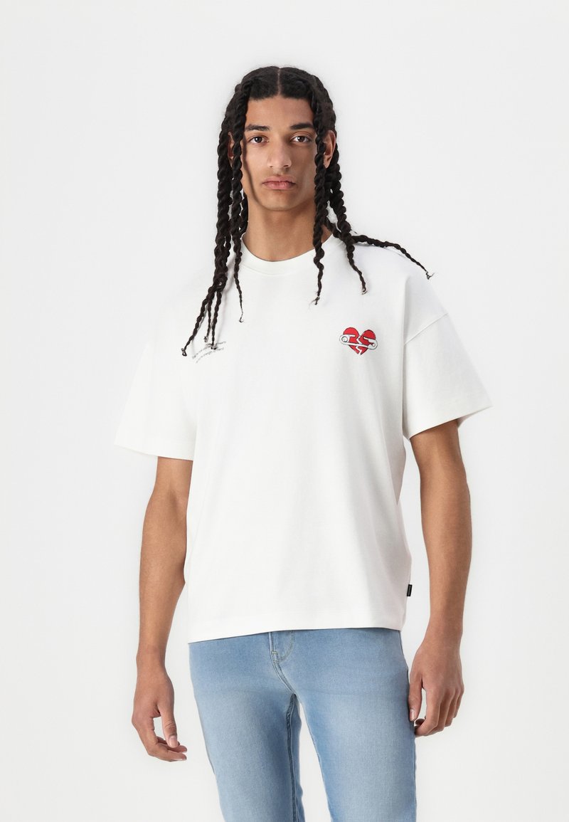 White cotton t-shirt with short sleeves, featuring a red heart graphic on the chest and minimalist text detail. Worn with light blue jeans.