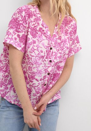 Kobieta w różowej bluzce na guziki z krótkim rękawem i kwiatowym wzorem oraz jasnoniebieskich dżinsach, stojąca z rękami złączonymi przed sobą.