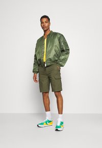 Grønn sateng bomberjakke, gul skjorte, oliven cargoshorts og grønne og gule joggesko. Modellen poserer med en hånd i lommen på shorts.