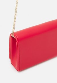 Punainen tekonahkainen clutch, jossa on suorakulmainen muoto, sileä pinta ja kultaketjuolkaimet. Sisältää näkyvää ompelutyötä sivuilla.