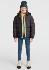 Svart puffjacka med huva, bärs över en grön fleece och en blå långärmad topp. Kombinerad med blå leggings och krämfärgade stövlar.