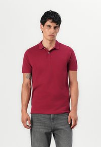 Mand med mørkt hår iført en rød kortærmet polo-shirt og grå jeans, stående mod en ensfarvet lys baggrund.