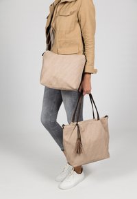 Beige fabric bag with a textured surface, featuring a long strap and tassel detail. The bag is spacious and rectangular in shape.