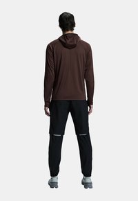 Felpa marrone a maniche lunghe con cappuccio, abbinata a pantaloni neri con accenti riflettenti. Il materiale sembra leggero e liscio.