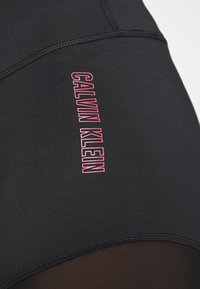 Mustat urheiluleggingsit, joissa on vaaleanpunainen "CALVIN KLEIN" -logo, sileä pinta ja kontrastivärinen verkkokangasdetaili alareidessä.