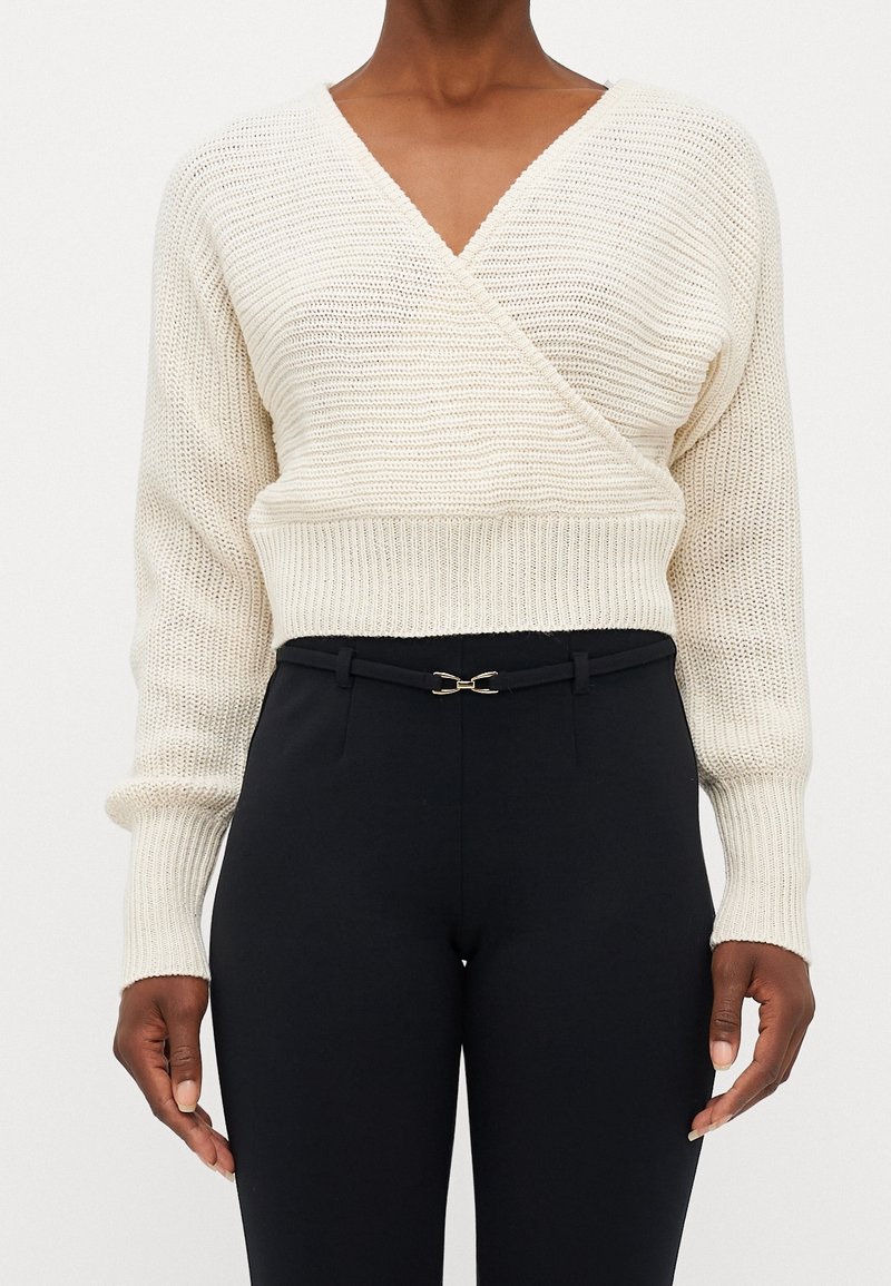 Personne à la peau foncée portant un pull en maille crème cache-cœur et un pantalon noir taille haute avec une ceinture fine noire et une boucle dorée.
