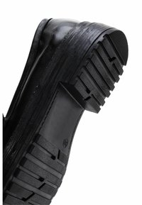 Zapato de goma negro con una suela gruesa y texturizada, que presenta profundos tacos y un empeine liso. Área de los dedos reforzada con un detalle mínimo.