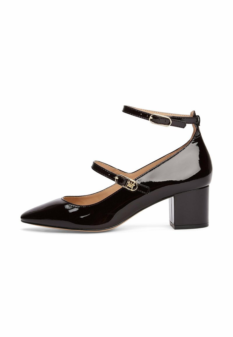 Chaussure à talon moyen en cuir verni noir avec bout pointu, doubles brides à la cheville et fermetures à boucle dorées sur fond blanc.