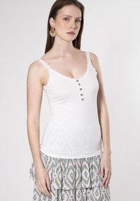Top senza maniche bianco con bordo in pizzo, caratterizzato da una chiusura a bottoni. Abbinato a una gonna verde e rosa a strati con motivo.