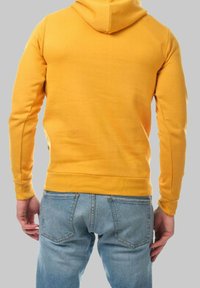 Hoodie jaune en tissu doux, doté d'une poche kangourou, d'une capuche fixée et de poignets côtelés. Associé à un jean bleu clair.