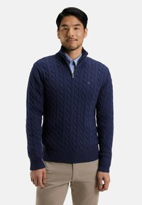 Pull en laine bleu marine avec un col zippé à un quart, des poignets et un ourlet côtelés. Porté par-dessus une chemise à col bleu clair.