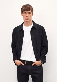 Mustat mokkajakku, jossa on kauluksellinen kaula, nappisuljenta ja pitkät hihat, päällä valkoinen t-paita, tummat farkut ja vyö.