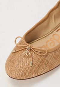 Beige ballet flats with a woven texture, rounded toe, rubber sole, and a decorative bow featuring metal accents and a small logo charm.