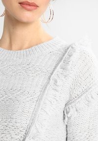 Zbliżenie na kobietę w białym, teksturowanym swetrze z frędzlami oraz dużymi kolczykami w kształcie kół, na jasnym tle.