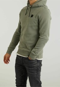Felpa verde in cotone con tasca frontale, cordini neri e un piccolo logo sul petto. Indossata con jeans scuri.