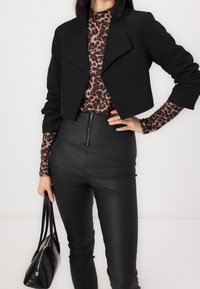 Černý krátký blazer přes hnědý top s leopardím potiskem, spárovaný s černými kalhotami s vysokým pasem, které mají přední zip a hladkou, jemnou texturu.