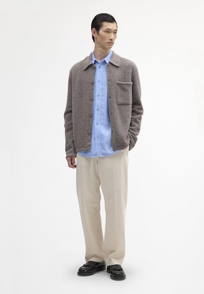 Cardigan marron en mélange de laine avec col, poche avant et fermeture à boutons, superposé sur une chemise bleu clair boutonnée et un pantalon beige.