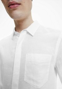 Camicia bianca in lino con colletto, dotata di taschino sul petto. Il tessuto ha un aspetto leggermente texturizzato. Branding discreto sulla tasca.