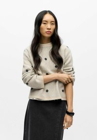 Beige sweater with black polka dots, textured knit, round neckline, and rolled cuffs, paired with a black denim skirt and a black wristband.