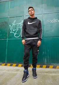 Felpa nera Nike e pantaloni jogger presentano un logo e dettagli bianchi, su uno sfondo verde. Sneakers alte nere e blu completano il look.