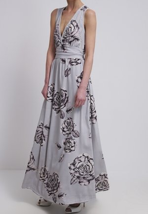 Sukienka maxi bez rękawów w jasnoszarym kolorze z głębokim dekoltem w kształcie litery V oraz dużymi czarno-białymi wzorami róż, noszona z białymi sandałami na obcasie z odkrytymi palcami.