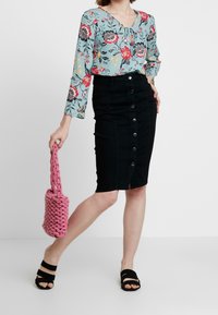 Bloemen blouse met lange mouwen in lichtblauw, zwarte denim kokerrok met knopen aan de voorkant, zwarte instapsandalen en een gestructureerde roze geweven handtas.