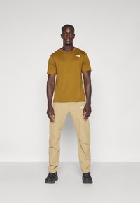 T-shirt de manga curta mostarda e calças bege, ambas com detalhes de logótipos. Ténis de desporto pretos completam o look. Textura plana, design casual.