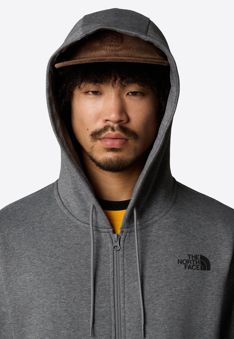 Sudadera gris con cremallera y un logo negro en el lado izquierdo del pecho, que cuenta con capucha con cordón y una textura de tela suave. Lleva encima una camiseta amarilla.