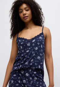 Camisole bleu marine à fleurs avec fines bretelles et bordure en dentelle. Présente une coupe décontractée et un tissu texturé avec de petits motifs de fleurs blanches.