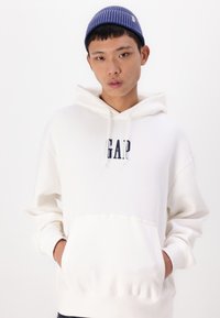 Sweat à capuche blanc avec une poche avant et le logo "GAP" en bleu foncé. Porté avec un bonnet rayé bleu et blanc. Texture lisse.