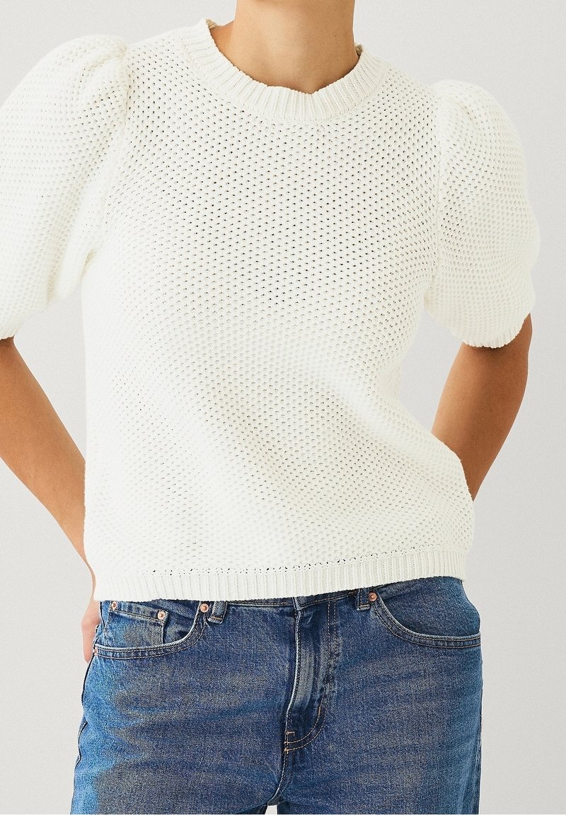 Maglione bianco a maniche corte lavorato a maglia con pattern testurizzato e colletto e orlo a coste. Abbinato a jeans in denim blu.