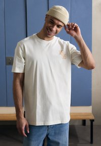 Krämfärgad bomullströja med korta ärmar, prydd med gyllene broderad text "1956" på bröstet och ihop med ljusblå jeans.