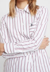 Chemise blanche à manches longues avec des rayures verticales rouges, dotée d'un petit logo brodé sur la poitrine et de poignets boutonnés.