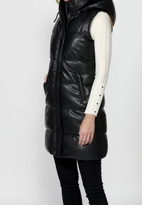 Femme portant un gilet en cuir matelassé noir sans manches par-dessus un haut blanc côtelé à manches longues avec des détails de boutons sur la manche.