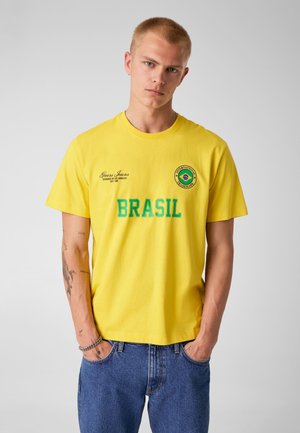 Giovane uomo con capelli biondi corti che indossa una t-shirt gialla con la scritta "BRASIL" e un emblema circolare della bandiera brasiliana, abbinata a jeans blu.