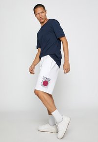 Camiseta de manga corta azul marino combinada con pantalones cortos blancos que presentan un gráfico rojo y azul de "Tokio"; zapatillas blancas y calcetines tobilleros.