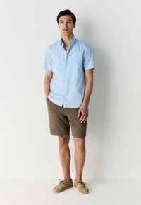 Lyseblå kortærmet skjorte med knaplukning ved kraven og brystlomme. Kombineret med brune shorts og lyse flettede snøresko.