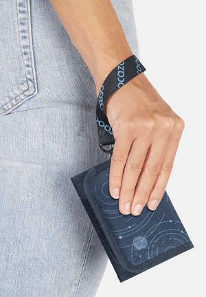 Hand holding navy blue patterned wallet with black wrist strap, wearing light blue denim jeans.