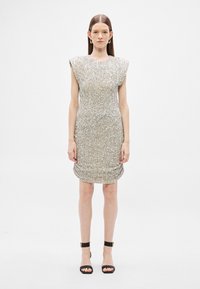 Vestido de lantejoulas prateadas com mangas curtas e estruturadas e design franzido. Usado com sandálias pretas com tiras. Fundo branco liso.